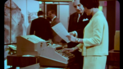 A woman in a suit operates a cash register at a booth at EuroShop 1968, while a man next to her shows a brochure; copyright: Messe Düsseldorf 