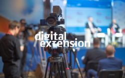 Eine Filmkamera im Vordergrund, eine Podiumsdiskussion mit Zuschauern im Hintergrund, darau weiß die Begriffe "Digital Extension"; copyright: Messe Düsseldorf/ctillmann