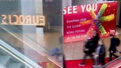 People walk past a large poster advertising EuroShop 2026; to the left, “EuroCIS” is reflected in neon lettering; Copyright: Messe Düsseldorf/A. Wiese 