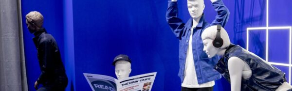 Shop window display with several mannequins in modern clothing against a blue-lit background, including one figure with a newspaper and one with headphones; copyright: Messe Düsseldorf/A. Wiese