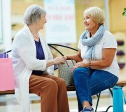 Two elderly women are sitting on a bench in a shopping mall, smiling and chatting, with several colorful shopping bags next to them.