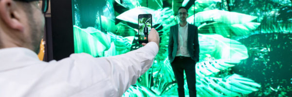 Image: Trade fair visitor is photographed by another visitor in front of a greenly lit wall; Copyright: Messe Düsseldorf/A. Wiese