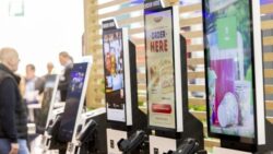 Several self-service ordering kiosks with touchscreens at a trade show booth; trade show visitors walking by in the background
