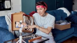 An Asian-looking person with a red headband sits on a sofa and presents a pair of sneakers in an open shoe box in front of a smartphone on a tripod.