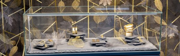 Gold jewelry in a decorated glass display case, arranged on dark stone slabs in front of a decorative wall with golden leaf motifs; copyright: Messe Düsseldorf/A. Wiese