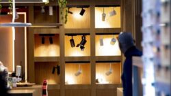 Exhibition area with illuminated wooden shelves displaying various spotlights and ceiling lights in different colors and lighting moods; on the right in the foreground, a… 