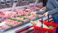 A woman with a shopping basket is pointing at something at the meat counter