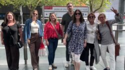 Seven members of the EuroShop team are posing together for a photo in front of the bus stop sign at Messe Düsseldorf. 