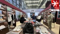 A modern store with wooden shelves full of clothing, a table with sneakers and accessories at the front, and the EuroShop on Tour logo in the top right corner. 