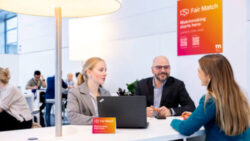 Three people are talking at EuroShop. In the background, a sign with the words Fair Match can be seen. 