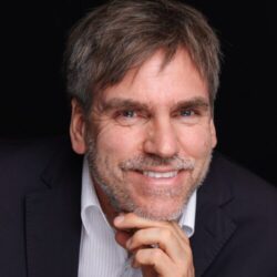 A friendly, smiling middle-aged man in a suit and white shirt, resting his head on his hand against a dark background.
