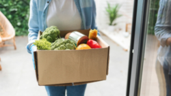 A person hands over a box of fresh groceries, including broccoli, bell peppers, and canned goods, at the doorstep as part of a supermarket delivery.