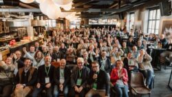 A large group of conference participants sits close together in a modern event space, looking at the camera and laughingly raising their hands. 