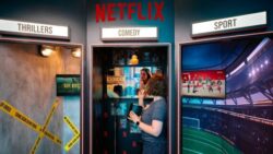 A person stands with popcorn in a Netflix installation between the “Thrillers,” “Comedy,” and “Sports” sections, looking at screens displaying various streaming content.