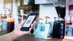 A person scans a QR code display at a café counter with a smartphone for mobile payment.
