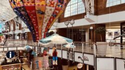 A colorful, decorative hot air balloon hangs from the ceiling of a modern shopping center with an open, bright upper floor and large windows. Below it, there is an artfully designed. 