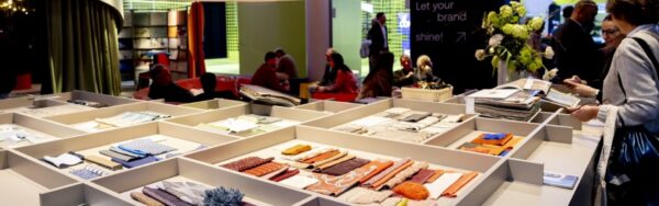 Exhibition table with fabric samples and textiles in various colors and textures, with exhibitors and visitors seated behind it engaged in conversation; copyright: Messe Düsseldorf/A. Wiese