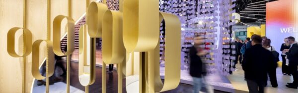 Designer exhibition stand with gold-colored, sculptural metal forms in front of light wood surfaces and an installation of hanging elements as visitors walk by; copyright: Messe Düsseldorf/A. Wiese