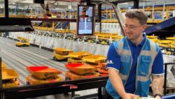 A GLS employee stands in an automated parcel sorting facility, wearing blue work clothing and a yellow safety vest. Behind him, numerous yellow containers move along a conveyor belt system.